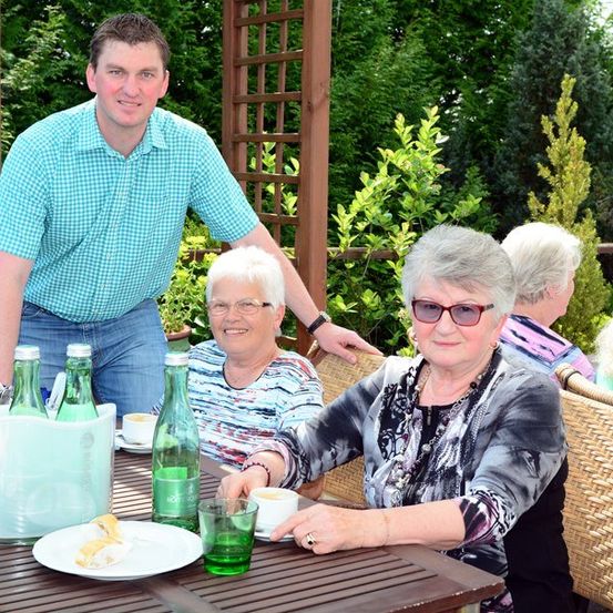 Eine Gruppe älterer Frauen und ein Mann sitzen um einen Tisch in einem Garten, lächeln und genießen ihre Getränke und das Essen.