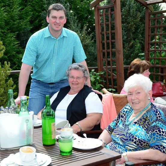 Eine Gruppe älterer Frauen und ein Mann sitzen an einem Tisch im Garten, lächeln und posieren für ein Foto. Auf dem Tisch stehen grüne Flaschen, Teller und Tassen. Dahinter befinden sich Holzgittern und Bäume.