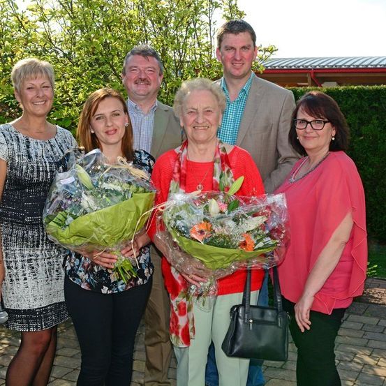 Eine Gruppe von Menschen, darunter zwei Frauen mit Blumensträußen, steht in einem Garten und lächelt für ein Foto.