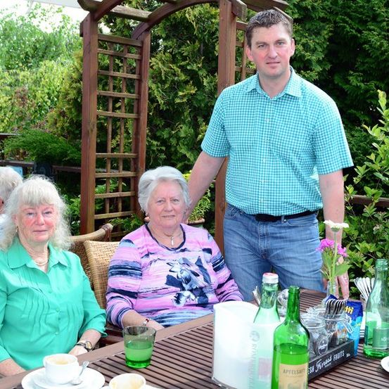 Drei Menschen sitzen an einem Tisch im Garten. Ein Mann steht hinter dem Tisch, und eine Frau sitzt links. Eine Frau in der Mitte hält eine Blume. Flaschen, Tassen und Teller stehen auf dem Tisch.