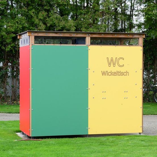 Eine bunte öffentliche Toilettenkabine mit 'WC' und 'Wicklelttisch' darauf, in einem Grasbereich mit Bäumen im Hintergrund.