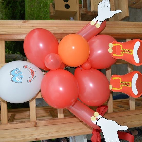 Eine Ballonfigur, die einem Clown ähnelt, mit roten und orangefarbenen Ballons, einem weißen Ballon mit Gesicht und einem roten Band. Die Figur steht auf einer Holzoberfläche.
