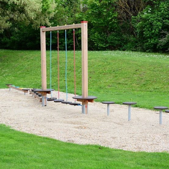 Ein Outdoor-Spielplatz mit einem Holzgerüst, Schaukeln und einem sandigen Untergrund, umgeben von saftigem grünem Gras und Bäumen.
