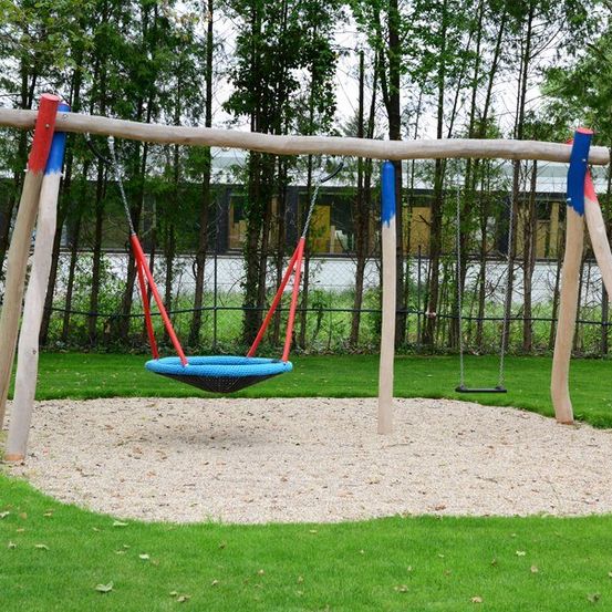 Ein Holzschaukelset mit einer blauen Schaukel und einer roten Schaukel, umgeben von grünem Gras und Bäumen.