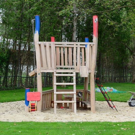Ein hölzernes Spielhaus mit einer Leiter und einer Rutsche, in einem sandigen Bereich, umgeben von grünem Gras und Bäumen. Ein Schubkarren ist in der Nähe.