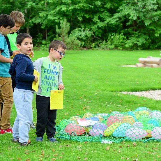 Eine Gruppe von Kindern steht auf einem Grasfeld, einige halten gelbe Papiere, und ein Haufen bunter Luftballons liegt auf dem Boden. Bäume und ein kleines Kind sind im Hintergrund.