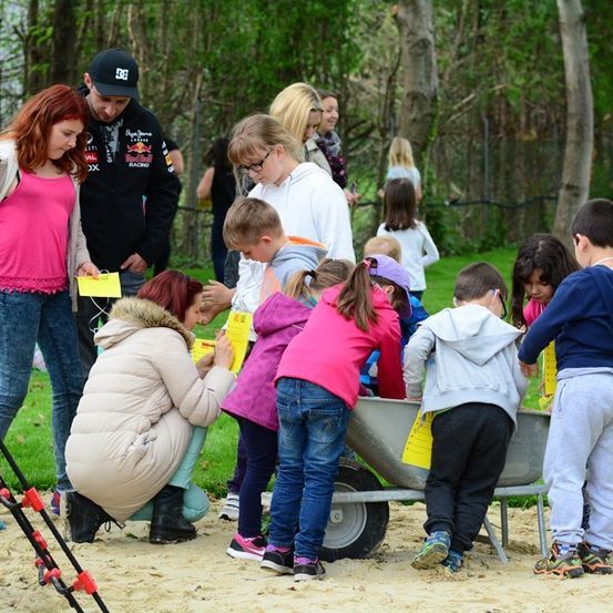 Eine Gruppe von Kindern und Erwachsenen versammelt sich um eine Schubkarre in einem Park, wobei einige Kinder gelbe Papiere halten. Die Erwachsenen helfen den Kindern, und im Hintergrund befinden sich Bäume und ein Zaun.