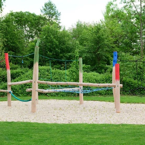 Ein Outdoor-Spielplatz mit Holzpfosten, bunten Seilen und einem Netz in einem von Bäumen umgebenen Grasbereich.