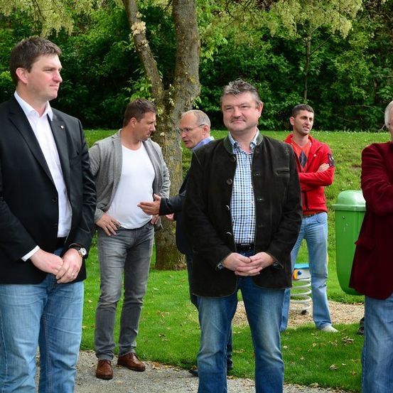 Eine Gruppe von Männern in Freizeitkleidung steht in einem Park, wobei einige in Gespräche verwickelt sind und andere beobachten. Die Umgebung umfasst üppige Grünflächen und einen Baumstamm.
