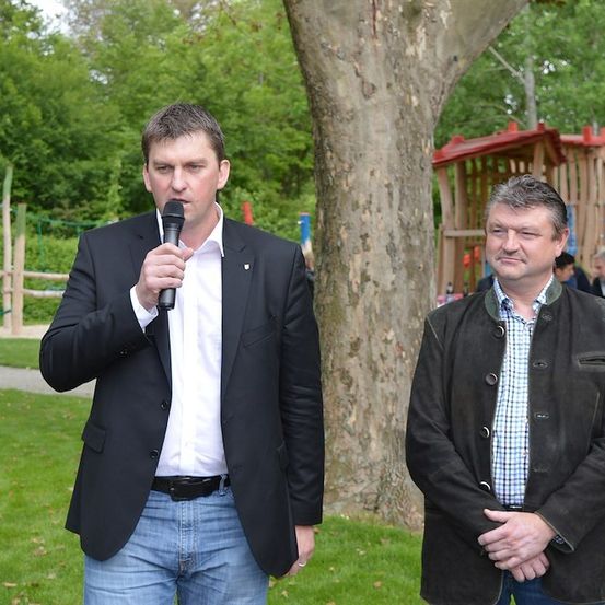 Zwei Männer stehen in einem Park, einer hält ein Mikrofon und spricht möglicherweise. Hinter ihnen ist ein Baumstamm und ein Spielplatz zu sehen.