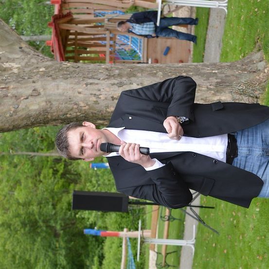 Ein Mann in einem schwarzen Anzug und Jeans hält ein Mikrofon und singt in einem Park. Hinter ihm sind ein Lautsprecher und ein Baumstamm zu sehen.