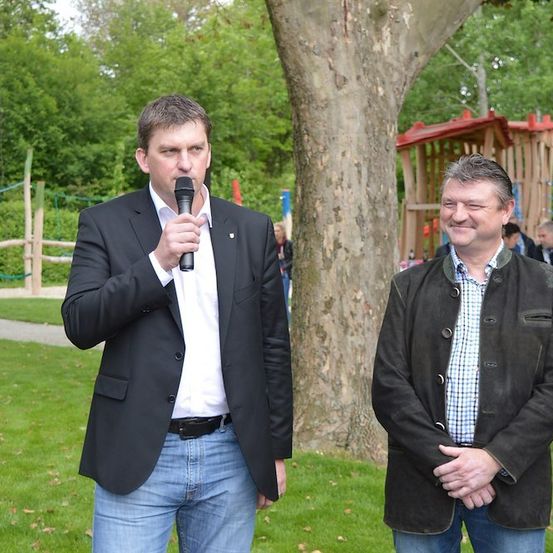 Zwei Männer stehen in einem Park, einer hält ein Mikrofon und spricht, während der andere zuhört. Hinter ihnen steht ein Baum und ein Spielplatz.