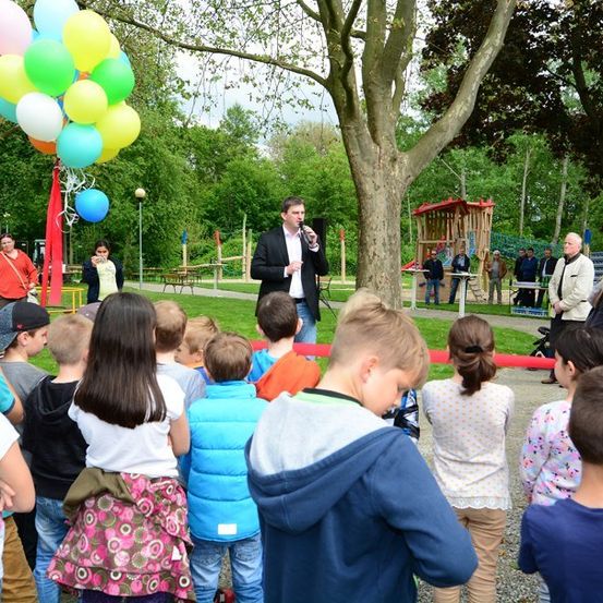 Ein Mann steht mit einem Mikrofon auf einer Bühne, umgeben von Luftballons, und spricht eine Gruppe von Kindern in einem Park an.