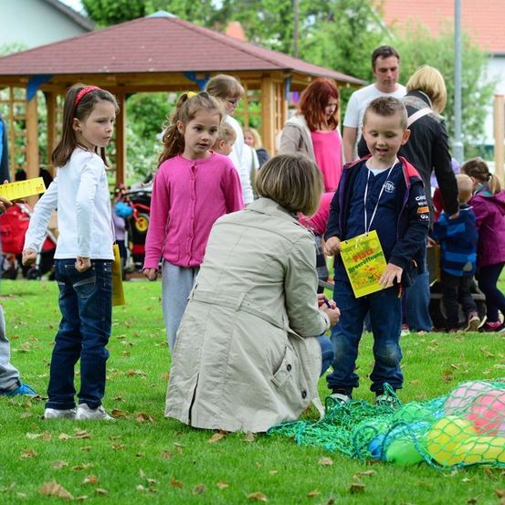 Eine Gruppe von Kindern und Erwachsenen versammelt sich in einem Park. Eine Frau kniet sich hin und interagiert mit einem Kind, das eine gelbe Karte hält. Bunte Luftballons sind auf dem Gras verstreut.