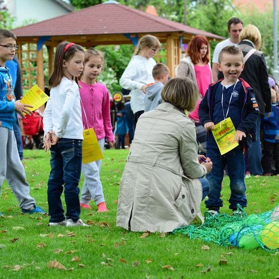 Eine Gruppe von Kindern und Erwachsenen steht auf einem Rasen. Einige Kinder halten gelbe Papiere. Eine Frau kniet und interagiert mit einem Kind. Ein Netz mit bunten Luftballons liegt auf dem Boden. Im Hintergrund steht ein hölzerner Pavillon und mehrere Menschen stehen.