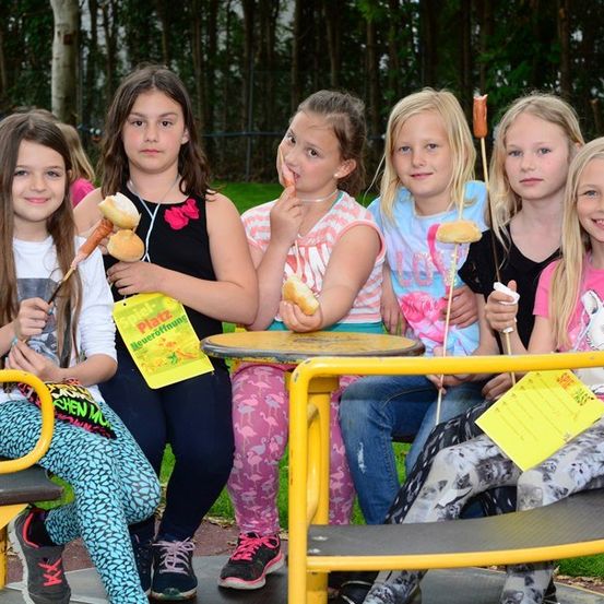 Sechs Mädchen sitzen auf einer gelben Bank in einem Park, jedes hält einen Hot Dog am Stiel und lächelt. Sie tragen Freizeitkleidung und scheinen eine Mahlzeit gemeinsam zu genießen.