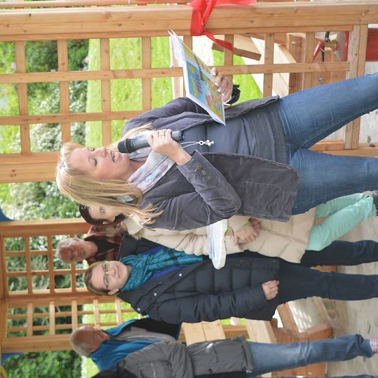 Eine Frau hält ein Buch und ein Mikrofon und singt zu einer Gruppe von Menschen in einem hölzernen Pavillon.