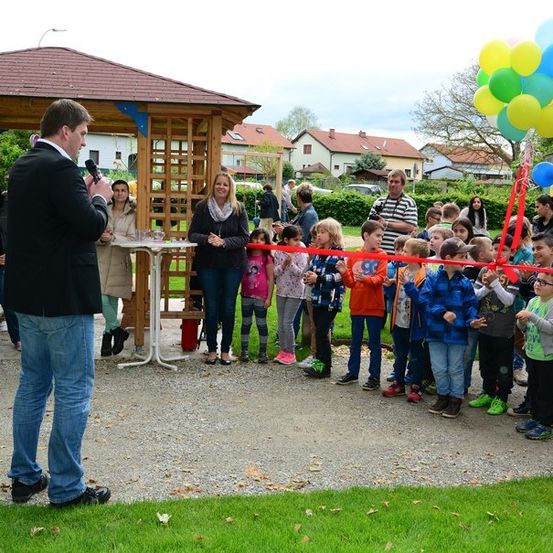 Ein Mann spricht in ein Mikrofon, während eine Gruppe von Kindern und Erwachsenen sich um eine Schleifenschneidezeremonie mit Luftballons und einem Pavillon im Hintergrund versammelt.