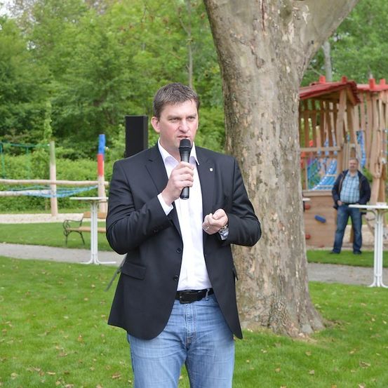 Ein Mann in einem schwarzen Anzug und Jeans spricht in ein Mikrofon in einem Park mit einem Baum und einem Spielplatz im Hintergrund.