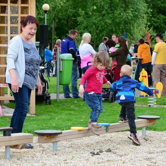 Eine Frau und zwei Kinder spielen auf einer Wippe in einem Park, während andere Familien im Hintergrund sind.