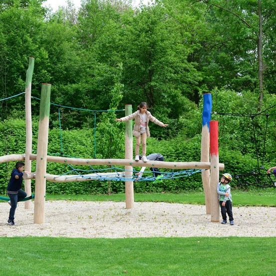 Eine Gruppe von Kindern spielt auf einem hölzernen Hindernisparcours in einem Park. Ein Kind balanciert auf einer Stange, während andere zuschauen.
