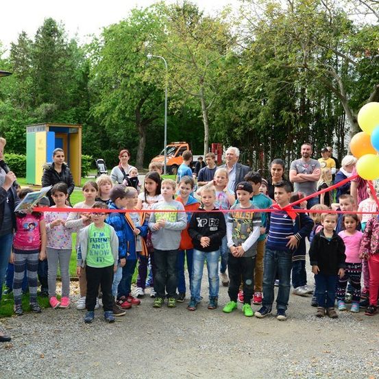 Eine Gruppe von Kindern und Erwachsenen steht in einer Reihe, hält ein rotes Band und eine Frau hält ein Buch und ein Mikrofon. Sie sind zu einer Schleifenschneide-Zeremonie versammelt.