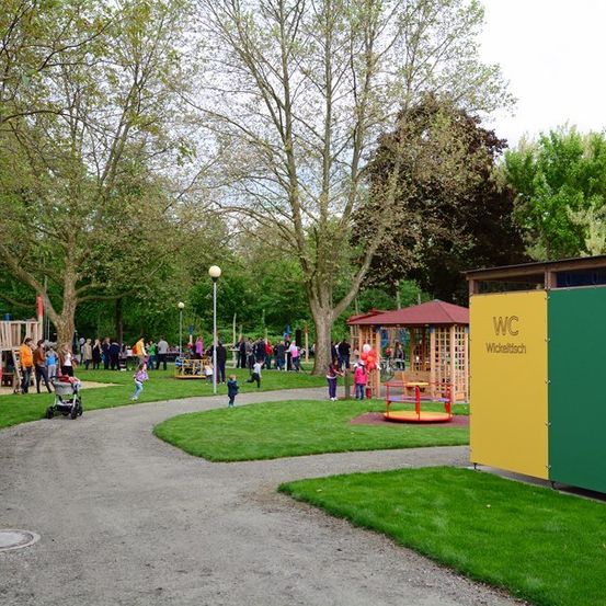 Ein Park mit einem Spielplatz, einem bunten WC-Schild und Menschen, die an einem sonnigen Tag herumlaufen.