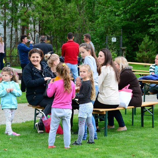 Eine Gruppe von Menschen, einschließlich Erwachsener und Kinder, sitzen auf Bänken in einem Park. Einige essen, während andere stehen und plaudern. Der Park ist von Bäumen und Gras umgeben.