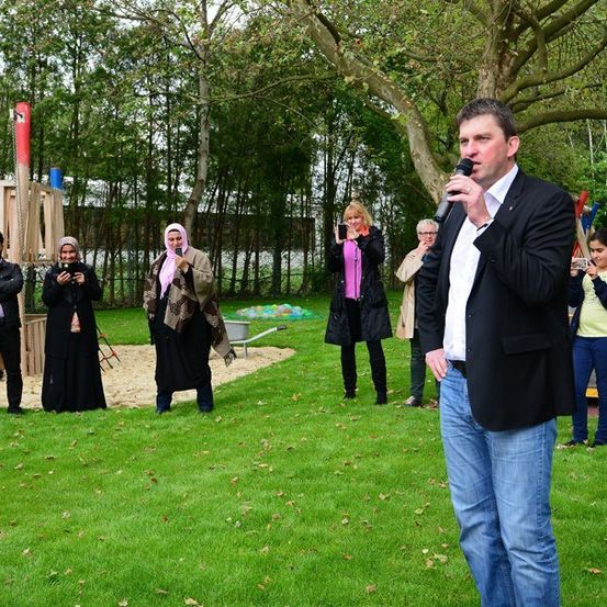 Ein Mann spricht in ein Mikrofon in einem Park, während mehrere Personen hinter ihm stehen und Fotos machen.
