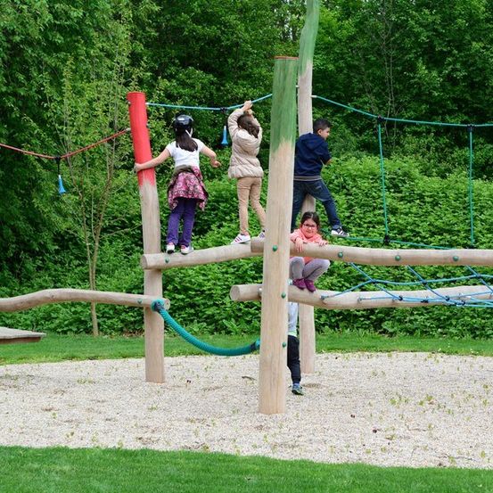 Eine Gruppe von Kindern spielt auf einem hölzernen Hindernisparcours in einem Park mit Grünpflanzen im Hintergrund.