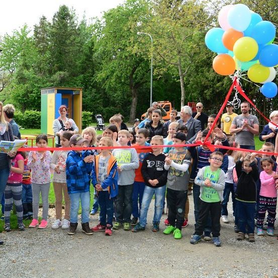 Eine Gruppe von Kindern und Erwachsenen steht bei einer Schleifenschneidezeremonie mit Luftballons und einer Frau, die ein Buch hält.