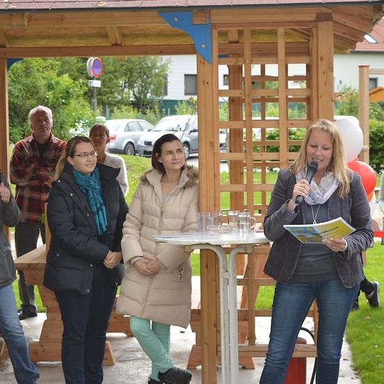 Eine Gruppe von Frauen und Männern steht in einem Pavillon, wobei eine Frau ein Mikrofon hält und spricht. Die Frau vorn hält ein Buch und ein Mikrofon. Es gibt Gläser und einen Tisch mit einem Buch darauf. In der Ferne sind Autos geparkt.