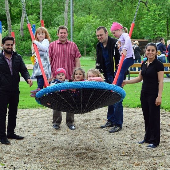 Eine Familie mit sechs Personen, darunter drei Kinder, steht um eine große Schaukel in einem Park mit grünem Rasen und Bäumen im Hintergrund.