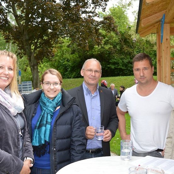 Fünf Menschen lächeln und posieren für ein Foto in einer Außenumgebung mit einem hölzernen Pavillon im Hintergrund. Sie stehen um einen Tisch mit Gläsern Wasser.