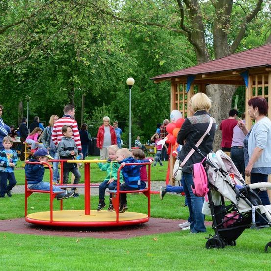 Mehrere Kinder spielen auf einem Karussell in einem Park, während Erwachsene und andere Kinder herumlaufen und zusehen. Im Hintergrund befindet sich ein hölzerner Pavillon.