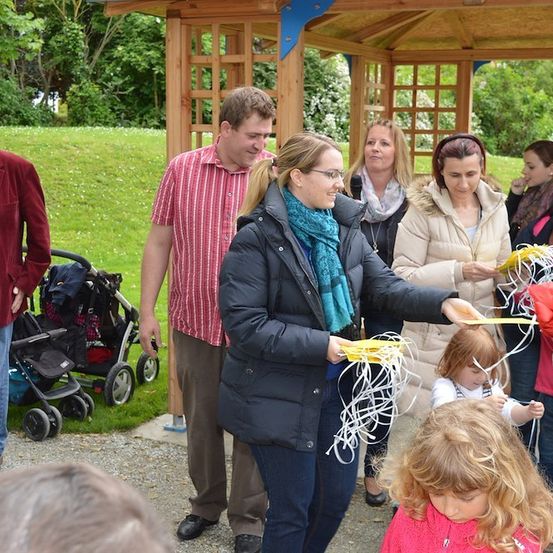 Eine Gruppe von Menschen, einschließlich Erwachsener und Kindern, versammelt sich in einem Park. Eine Frau mit einem blauen Schal hält eine Menge Bänder, und ein Kind hält ein Band. Sie stehen vor einem Pavillon.