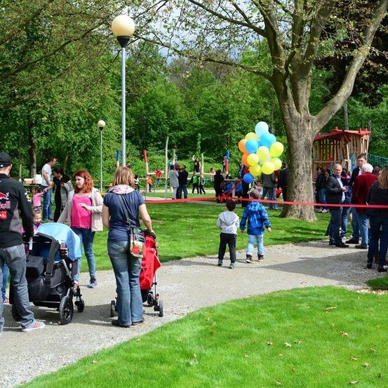 Eine Gruppe von Menschen versammelt sich in einem Park, einige gehen mit Kinderwagen, andere stehen herum. Ein Band ist über den Weg gespannt, und eine Person hält einen Strauß Luftballons. Eine Holzstruktur befindet sich auf der rechten Seite.