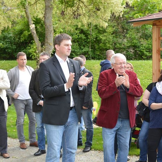 Eine Gruppe von Menschen, darunter ein Mann in einem schwarzen Anzug, der applaudiert, steht in einem Park mit grünem Rasen und Bäumen im Hintergrund.