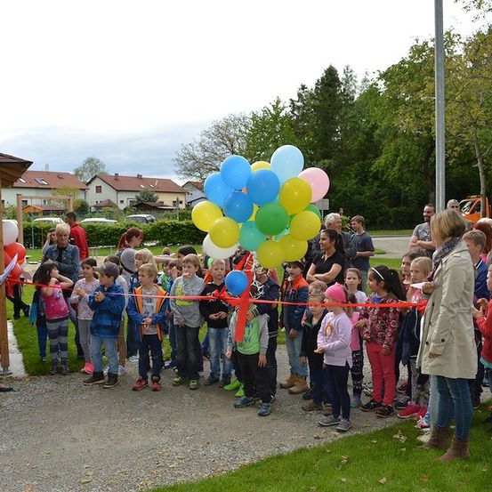Eine Gruppe von Kindern und Erwachsenen versammelt sich im Freien für eine Eröffnungsfeier, hält Luftballons und steht vor einem roten Band.
