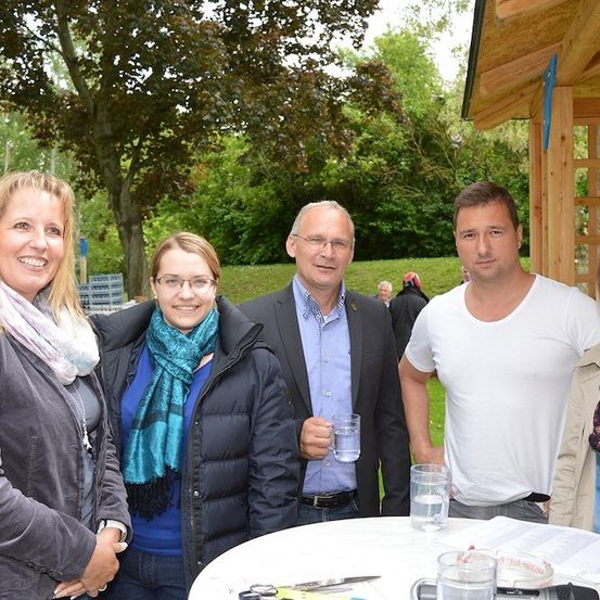 Fünf Erwachsene stehen vor einem hölzernen Pavillon und lächeln für ein Foto. Ein Mann im Anzug hält ein Glas Wasser. Eine Frau in einem blauen Hemd und Schal steht zwischen zwei Männern. Eine Frau in einer grauen Jacke steht links.