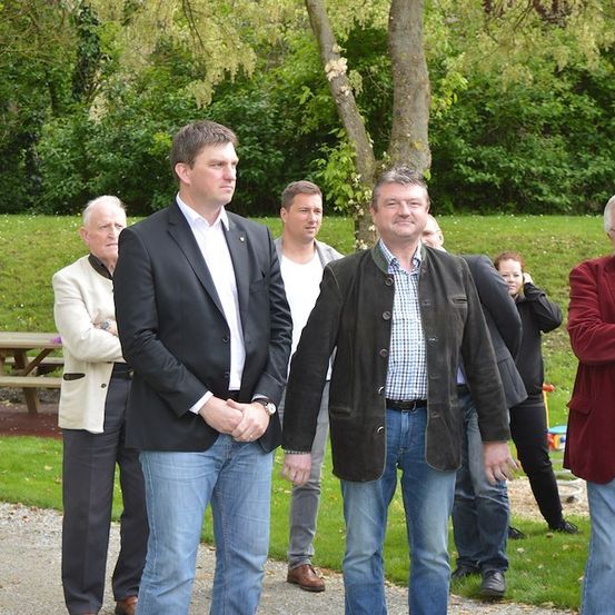 Eine Gruppe von Männern in formeller und lässiger Kleidung steht in einem Park, einer Mann trägt eine Brille und ein anderer hält eine Zigarette. Hinter ihnen ist ein Picknicktisch und ein Baum zu sehen.