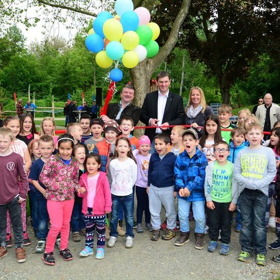 Eine Gruppe von Kindern und Erwachsenen steht in einem Park mit einem Baum und Luftballons und feiert ein Ereignis, bei dem ein roter Faden von zwei Männern in Anzügen geschnitten wird.