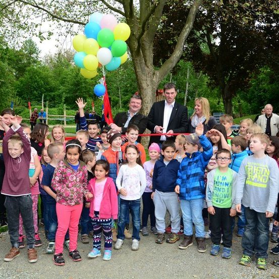 Eine Gruppe von Kindern und Erwachsenen steht in einem Park und feiert mit Luftballons und einer Schleifenschneidezeremonie.