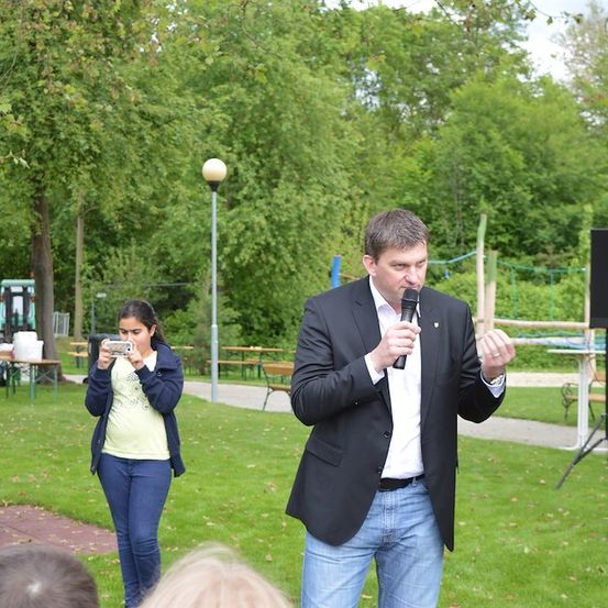 Ein Mann in einem Anzug spricht in ein Mikrofon in einem Park, während eine Frau ein Foto macht.