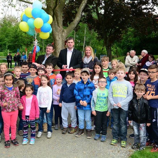 Eine Gruppe von Kindern und Erwachsenen versammelt sich um einen Baum mit Luftballons, möglicherweise für eine Eröffnungszeremonie. Die Erwachsenen stehen vorne, die Kinder dahinter. Das Gelände ist von Bäumen und Gras umgeben.