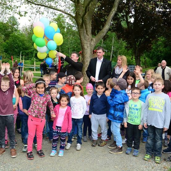 Eine Gruppe von Kindern und Erwachsenen versammelt sich in einem Park, einige halten Luftballons hoch. Ein Mann in einem Anzug steht vor ihnen.