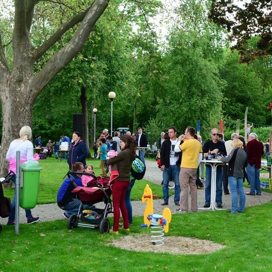 Eine Gruppe von Menschen versammelt sich in einem Park, einige stehen und andere sitzen um Tische herum. Eine Frau schiebt einen Kinderwagen mit einem Kind. Ein Baum und ein Spielplatz sind im Hintergrund.