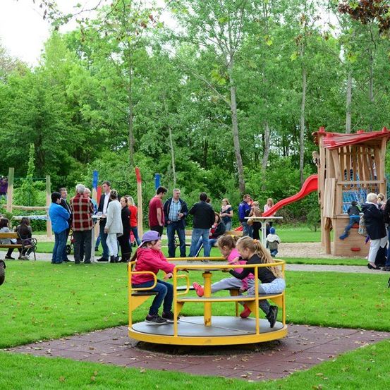 Eine Gruppe von Kindern und Erwachsenen spielt in einem Park mit verschiedenen Spielgeräten, darunter eine gelbe Drehscheibe und eine rote Rutsche.