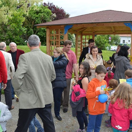 Eine Gruppe von Menschen, einschließlich Erwachsener und Kinder, versammelt sich um einen hölzernen Pavillon in einem Park, einige halten Luftballons und andere schauen sich etwas an.