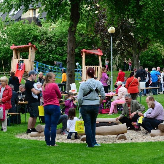 Eine Gruppe von Menschen versammelt sich in einem Park mit Spielplatz und Picknicktischen. Einige sitzen auf Bänken, andere stehen. Ein Mann hält ein Baby und eine Frau eine Tasche. Ein gelbes Schild liegt auf dem Boden.